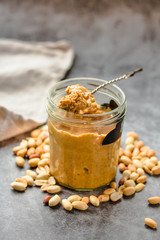 Fresh peanut spread in jar and spoon and scattered peanuts on gray background.