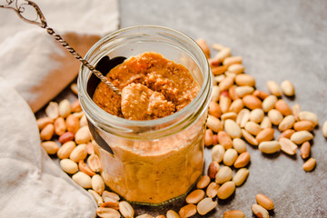Peanut paste in an open jar and and spoon, peanuts on the gray table.