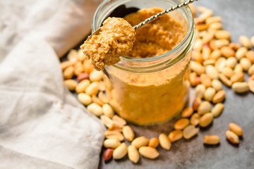 Fresh peanut spread in jar and spoon and scattered peanuts on gray background.Close-up.