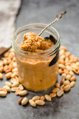 Fresh peanut spread in jar and spoon and scattered peanuts on gray background.