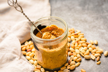 Peanut paste in an open jar and and spoon, peanuts on the gray table.Close-up.