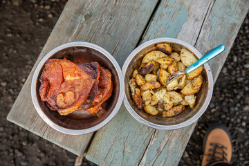 Metal bowls with lamb steaks and potatoes on wooden picnic table. Top view.