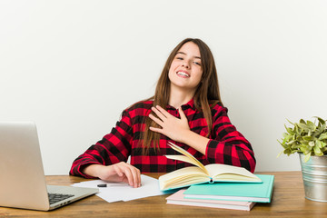 Young teenager going back to her routine doing homeworks laughs out loudly keeping hand on chest.