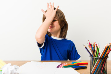 Little boy painting and doing homeworks on his desk forgetting something, slapping forehead with palm and closing eyes.