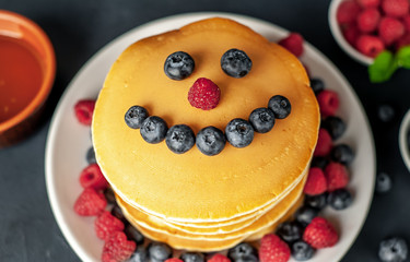American pancakes with raspberries, fresh blueberries and honey. Healthy breakfast on concrete background, funny face on pancakes painted with blueberries