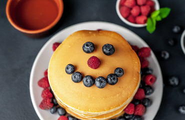 American pancakes with raspberries, fresh blueberries and honey. Healthy breakfast on concrete background, funny face on pancakes painted with blueberries