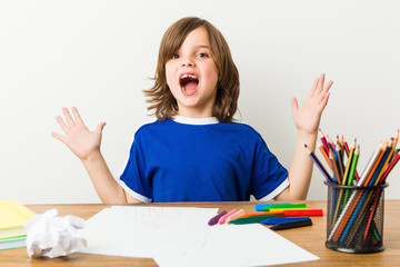 Little boy painting and doing homeworks on his desk celebrating a victory or success, he is surprised and shocked.