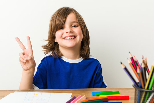 Little boy painting and doing homeworks on his desk joyful and carefree showing a peace symbol with fingers.