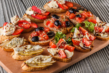 Assorted bruschetta with tomato, olives, camembert, blue cheese, lettuce, pear, honey on wooden board
