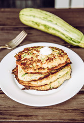 Fried zucchini pancakes stacked on plate, healthy organic summer food made with zucchini. Brown wood board background.