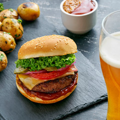 Delicious cheeseburger with baked potatoes and broccoli on a black stone board served with a glass of lager beer.