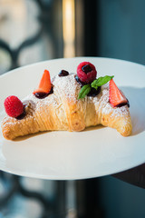 Croissant su piatto bianco con decorazioni di frutta, cioccolato e zucchero a velo