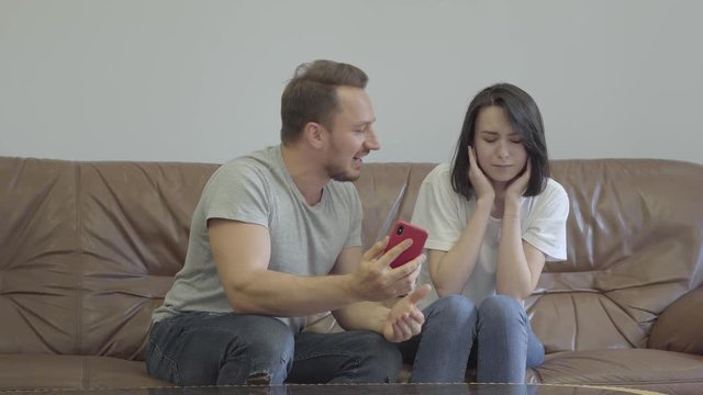 Portrait of angry man shouting at his wife showing her cellphone with messages from her paramour at home. Problems in the relationship between man and woman. Betrayal, mistrust, breakup concept.