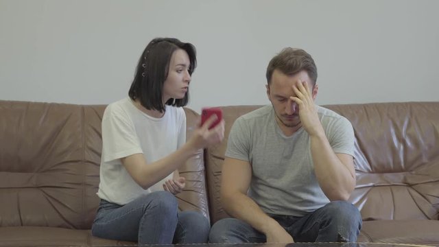 Portrait of angry woman shouting at her sad husband showing cellphone with messages from his paramour at home. Problems in the relationship between man and woman. Betrayal, mistrust, breakup concept