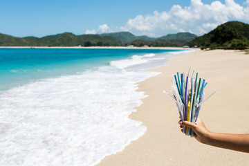 Hand holding heap of used plastic straws on background of clean beach and ocean waves. Plastic ocean pollution, environmental crisis. Say no plastic. Single-use plastic waste