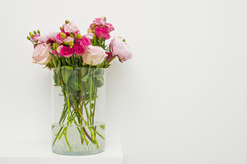 Bouquet of pink roses, peony flowers in glass vase on a shelf against white wall background.