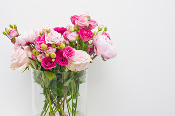 Bouquet of pink roses, peony flowers in glass vase on a shelf against white wall background.