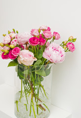 Bouquet of pink roses, peony flowers in glass vase on a shelf against white wall background.