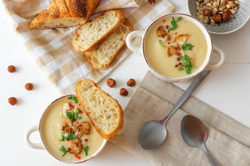 White wooden table with 2 bowls of cauliflower soup embellished with crushed hazelnuts, red pepper, smooth parsley herbs, Swiss twisted bread, frying pan with cauliflower, mortar and yellow flowers.