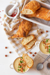 White wooden table with 2 bowls of cauliflower soup embellished with crushed hazelnuts, red pepper, smooth parsley herbs, Swiss twisted bread, frying pan with cauliflower, mortar and yellow flowers.