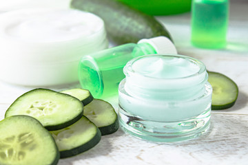 Obraz na płótnie Canvas Face cream in a glass jar with cucumber extract next to slices of fresh cucumbers on a white wooden background.