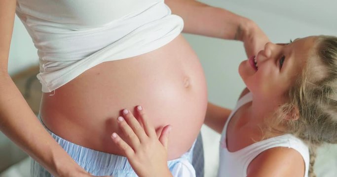 Authentic slow motion shot of a lovely little daughter is touching mother's pregnant belly, she is feeling baby at mother tummy and awaiting the birth of her little brother or sister.