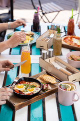 People are having breakfast on summer terrace: israeli shakshuka with tomatoes, salami and fried eggs, orange juice, omelette and light snacks