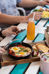 People are having breakfast on summer terrace: israeli shakshuka with tomatoes, salami and fried eggs, orange juice, omelette and light snacks