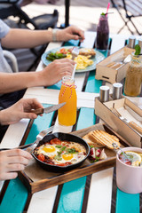People are having breakfast on summer terrace: israeli shakshuka with tomatoes, salami and fried eggs, orange juice, omelette and light snacks