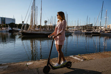 Young beautiful woman in a pink suit riding an electric scooter in the port of the sea,  modern girl, new generation, electric transport, ecology, ecological transport, dawn, electric skateboard