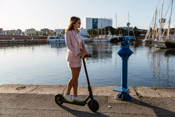 Young beautiful woman in a pink suit riding an electric scooter in the port of the sea,  modern girl, new generation, electric transport, ecology, ecological transport, dawn, electric skateboard