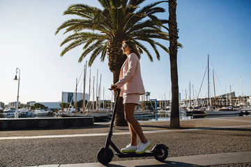 Young beautiful woman in a pink suit riding an electric scooter in the port of the sea,  modern girl, new generation, electric transport, ecology, ecological transport, dawn, electric skateboard