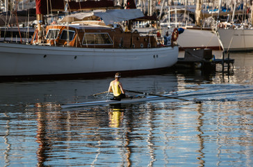 Obraz premium A man in a yellow shirt kayaking in a port near yachts and boats at dawn. Summer travel kayaking. Man paddling canoe. Exploring sea on vacation.
