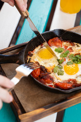 Woman is having breakfast on summer terrace: israeli shakshuka with tomatoes, salami, chili pepper and fried eggs, served in hot cast pan with toasts