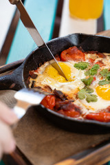 Woman is having breakfast on summer terrace: israeli shakshuka with tomatoes, salami, chili pepper and fried eggs, served in hot cast pan with toasts