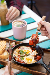 Woman is having breakfast on summer terrace: israeli shakshuka with tomatoes, salami, chili pepper and fried eggs, served in hot cast pan with toasts