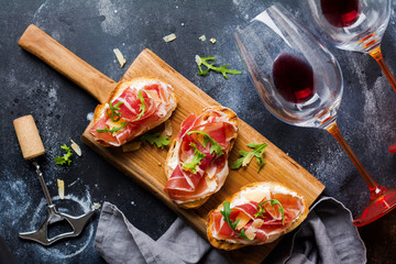 Open ham sandwiches, arugula and hard cheese, served on  wooden stand with aglass of red wine on aconcrete old dark background. Rustik style. Top view.