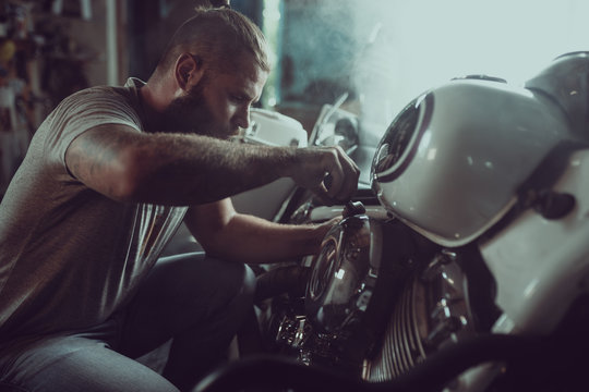 Handsome Bearded Man Repairing His Motorcycle In The Garage. A Man Wearing Jeans And A T-shirt