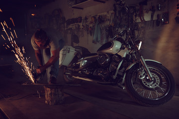 Obraz na płótnie Canvas Handsome brutal male with a beard repairing a motorcycle in his garage working with a circular saw. In the garage a lot of sparks and smoke from sawing