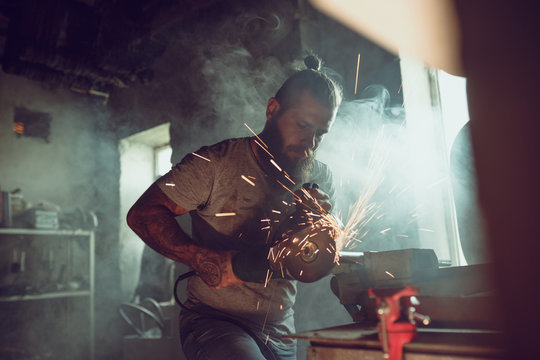 Handsome brutal male with a beard repairing a motorcycle in his garage working with a circular saw. In the garage a lot of sparks and smoke from sawing