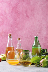 Variety of apple drinks. Bottles ang glasses of apple juice, vinegar and cider with garden apples with leaves and branches on table with pink background. Autumn home harvesting.