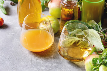 Variety of apple drinks. Bottles ang glasses of apple juice, vinegar and cider with garden apples with leaves and branches on grey texture table. Autumn home harvesting.
