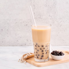 Tapioca pearl ball bubble milk tea, popular Taiwan drink, in drinking glass with straw on marble white table and wooden tray, close up, copy space.