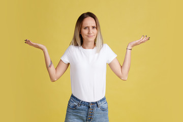 Young feminine woman does not understand and looking at camera in a white T-shirt and jeans stands on a yellow background. Advertising space