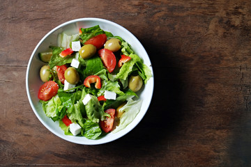 Traditional greek salad with fresh ingredients, feta cheese, olives, red tomatoes, cucumbers and greens in white ceramic bowl on wood textured background. Top view, close up, copy space.