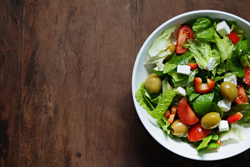 Traditional greek salad with fresh ingredients, feta cheese, olives, red tomatoes, cucumbers and greens in white ceramic bowl on wood textured background. Top view, close up, copy space.