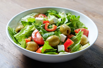 Traditional greek salad with fresh ingredients, feta cheese, olives, red tomatoes, cucumbers and greens in white ceramic bowl on wood textured background. Top view, close up, copy space.