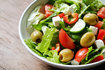 Traditional greek salad with fresh ingredients, feta cheese, olives, red tomatoes, cucumbers and greens in white ceramic bowl on wood textured background. Top view, close up, copy space.