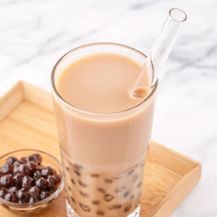 Tapioca pearl ball bubble milk tea, popular Taiwan drink, in drinking glass with straw on marble white table and wooden tray, close up, copy space.