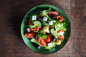 Traditional greek salad with fresh ingredients, feta cheese, olives, red tomatoes, cucumbers and greens in white ceramic bowl on wood textured background. Top view, close up, copy space.